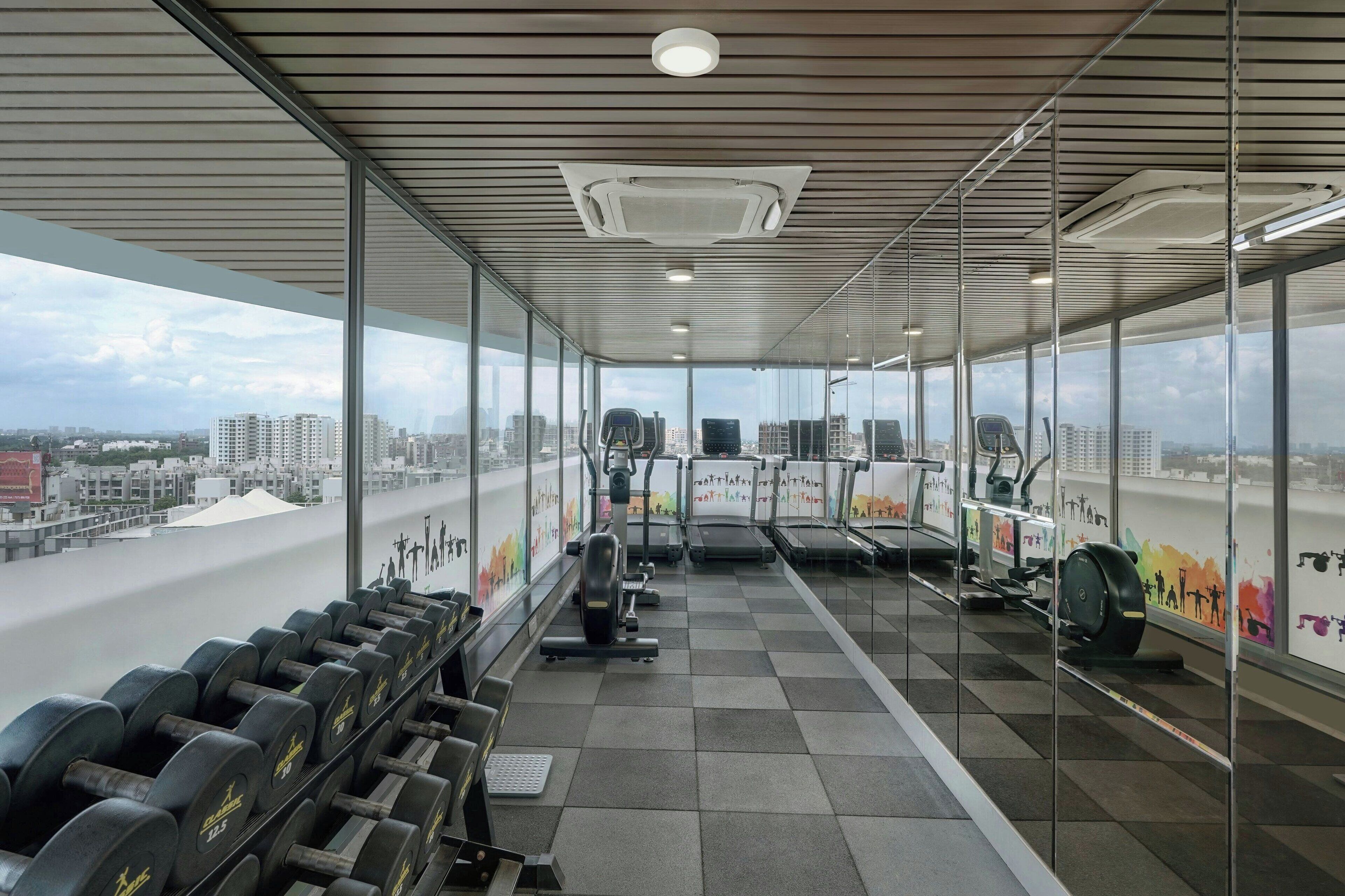 Une salle de sport équipée de matériel d’exercice, dotée de grandes fenêtres et offrant une vue sur la ville.