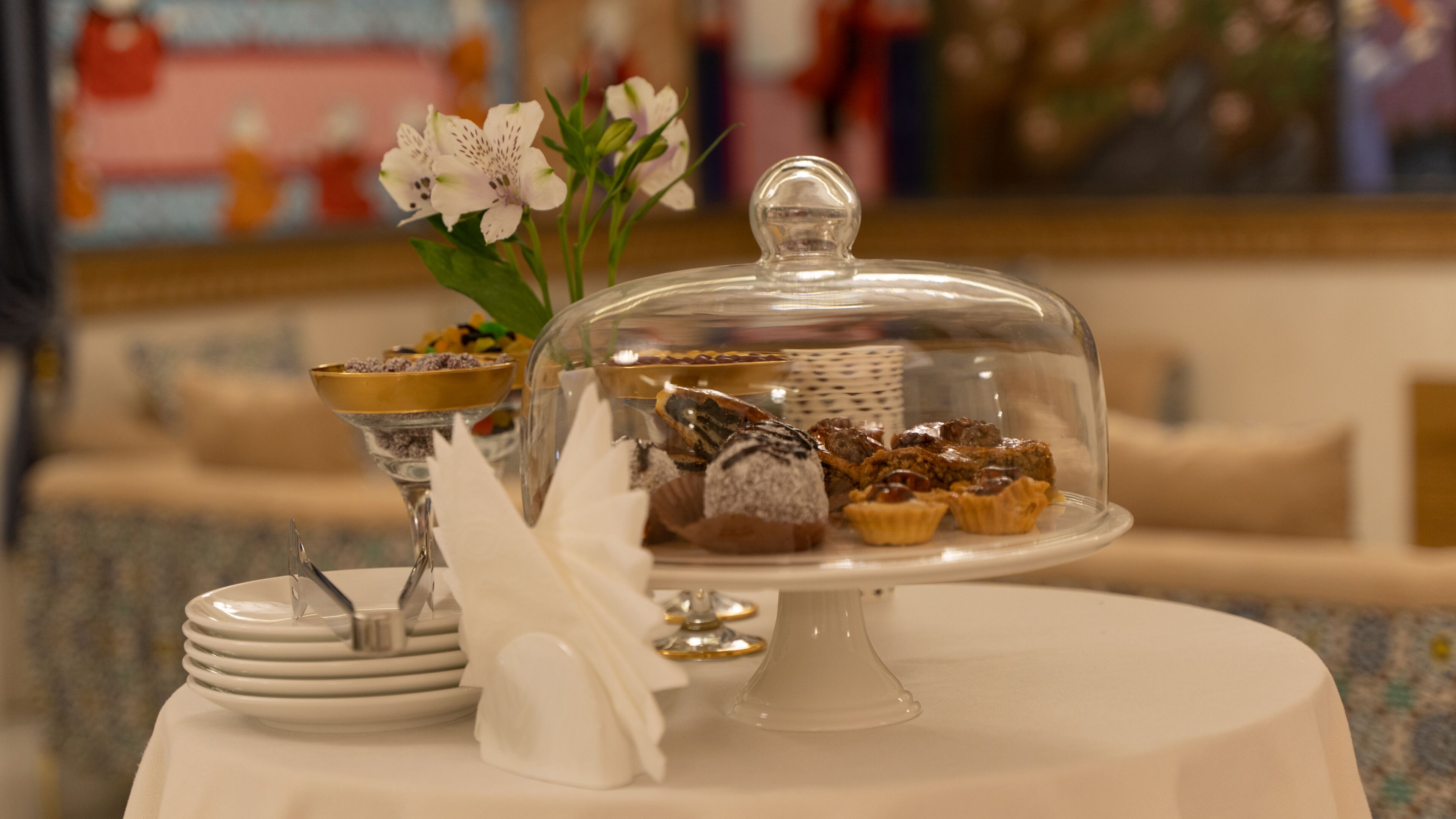 Una presentación de postres con una cubierta de vidrio abovedada, un tazón de dulces variados y un jarrón con flores sobre una mesa.