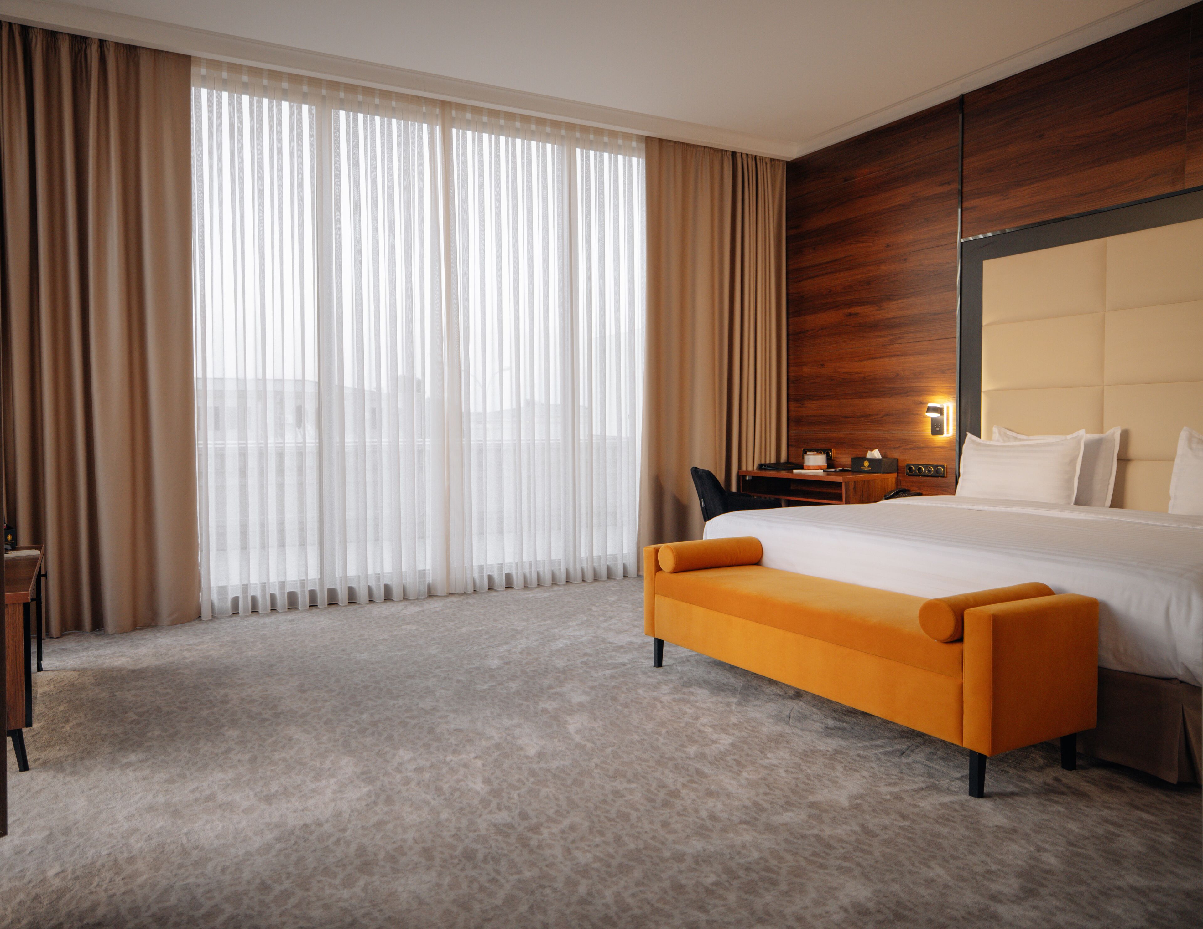 Habitación de hotel con una cama grande, cabecera de madera, escritorio con silla y un ventanal con cortinas.