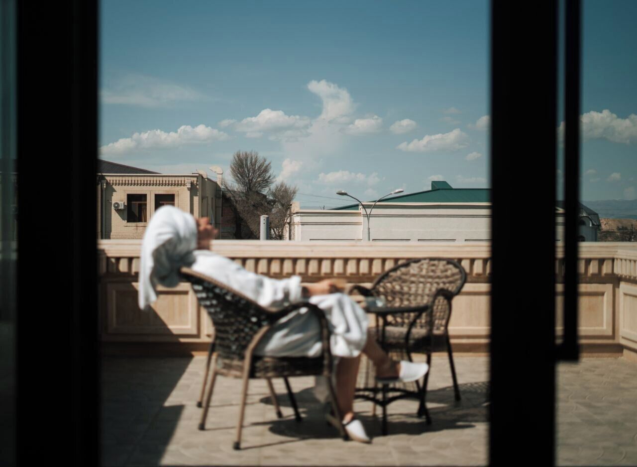 Un balcon avec une personne assise sur une chaise, une autre personne debout, et un bâtiment en arrière-plan.