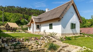 Une maison traditionnelle avec un toit de chaume, des murs blancs et des volets verts, entourée d’un jardin de pierres et d’un banc en bois.