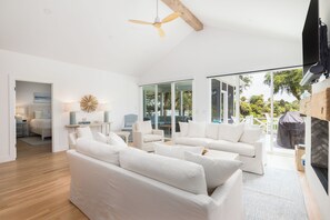A modern living room with white sofas, a ceiling fan, and a view of the outdoors through large glass doors. - Permanent Waves, Gorgeous home, Golf Course Views! (Isle Of Palms)