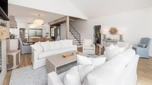 A spacious living area with a large white sofa, a wooden coffee table, and a ceiling fan. A staircase leads to an upper level, and there is a dining area visible in the background.