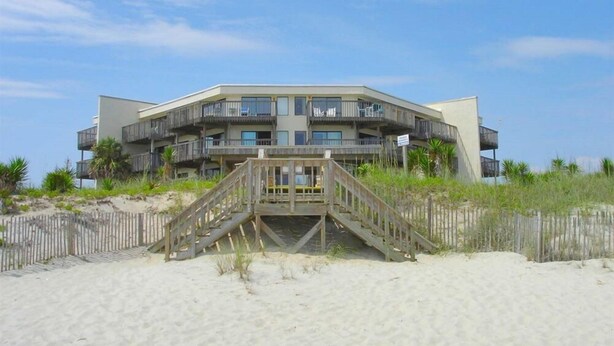 A beachfront building with multiple balconies, a wooden staircase leading to the entrance, and a sandy beach in the foreground.