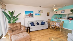 A living room with a sofa, ottoman, and a dining area with chairs and a table. There is a large plant, artwork on the walls, and a ceiling fan.