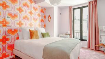 A bedroom with a bed, pillows, a bedside table, a window with curtains, and a wall with a floral pattern.