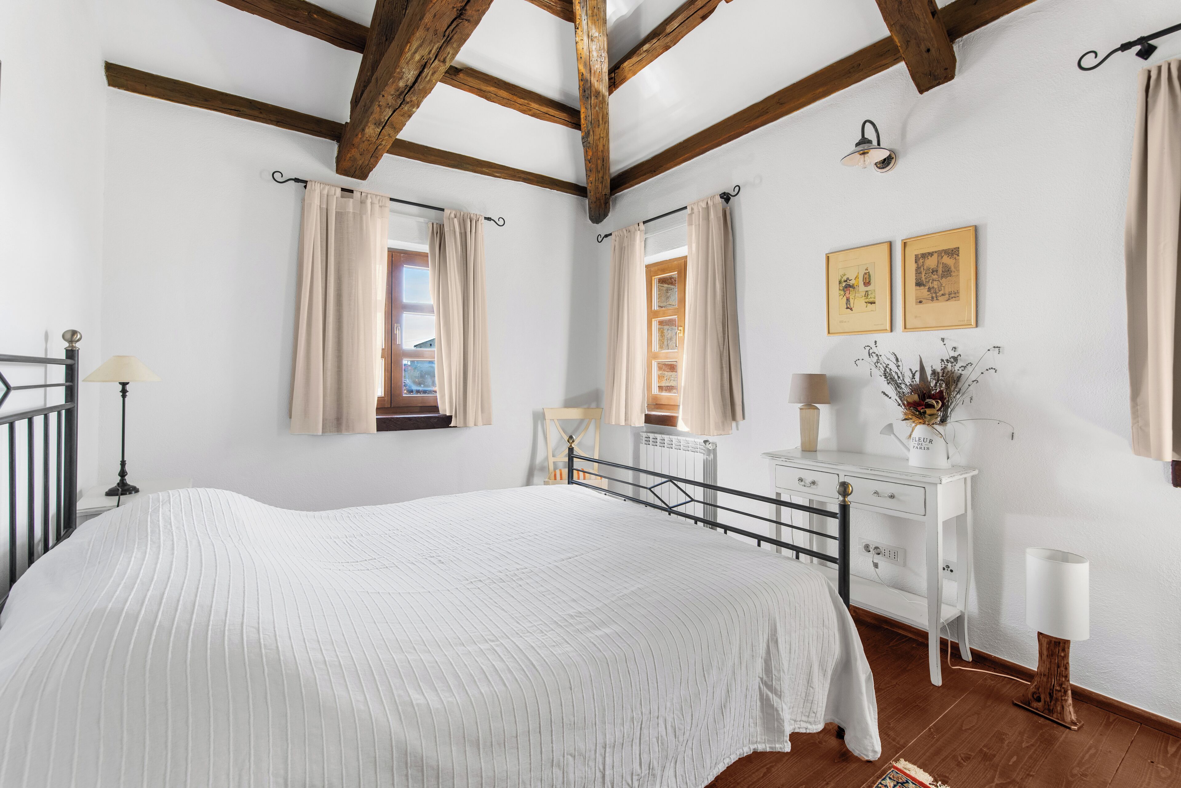 A bedroom with a large bed, wooden beams, a white dresser, and a window with curtains.