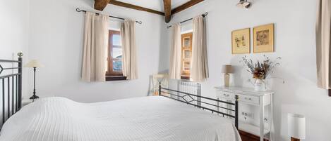 A bedroom with a large bed, wooden beams, a white dresser, and a window with curtains.