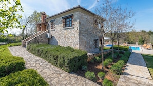 A stone house with a stone staircase leading to a wooden deck, surrounded by well-maintained hedges and a swimming pool.