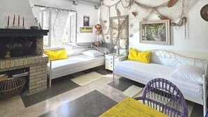 A compact room with a bed, a small table, a chair, a fireplace, and a window with blinds.