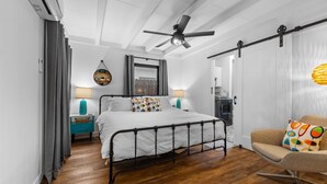 A bedroom with a metal bed frame, wooden flooring, a ceiling fan, and a view through a window.