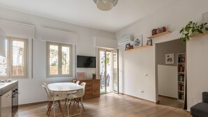 Un salón amplio con suelo de madera, una mesa de comedor con sillas, una zona de cocina con estufa y un televisor montado en la pared.