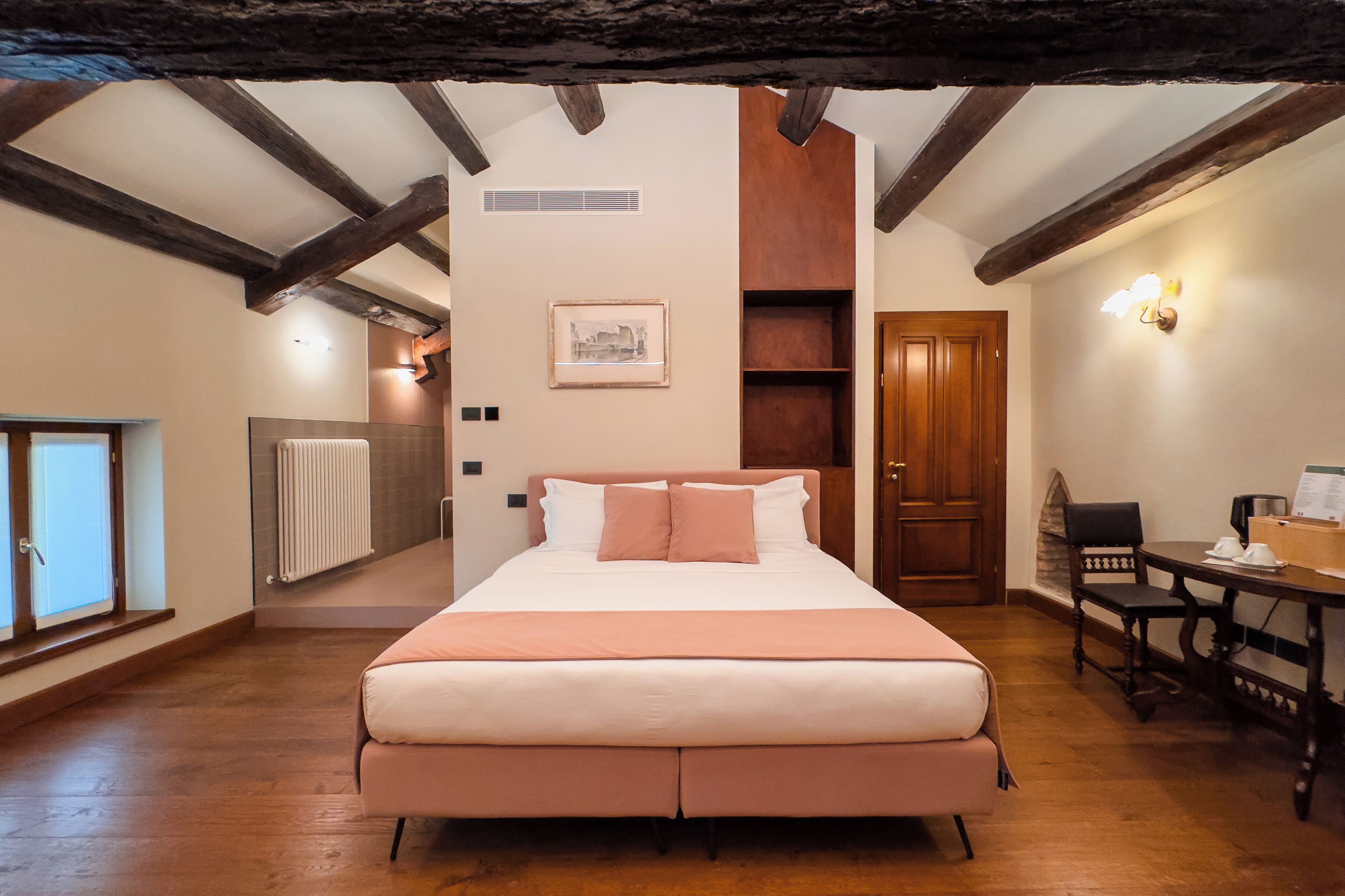 A bedroom with a large bed, wooden beams, a radiator, a framed picture, a wooden door, and a table with a chair.