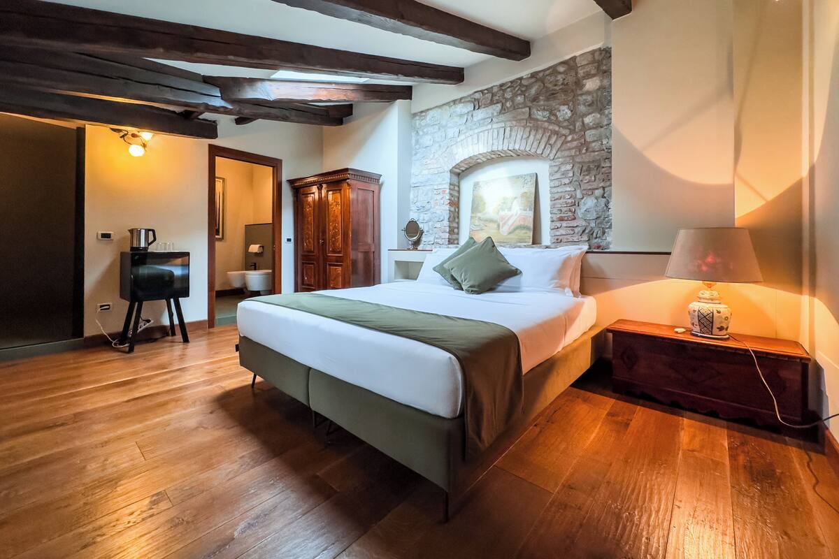 Un dormitorio con piso de madera, una cama con una colcha verde, una mesita de noche con lámpara, una pared de piedra y un armario de madera.