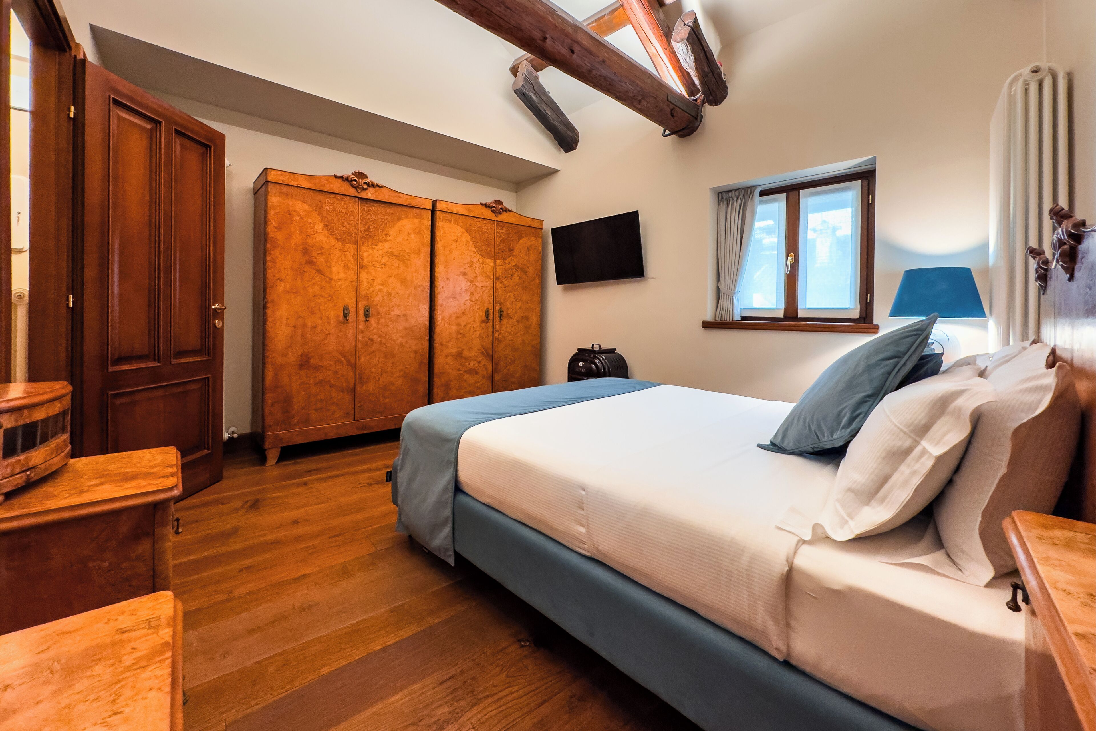 Quarto com guarda-roupa de madeira, cama com roupa de cama azul, janela com cortinas e televisão de tela plana na parede.
