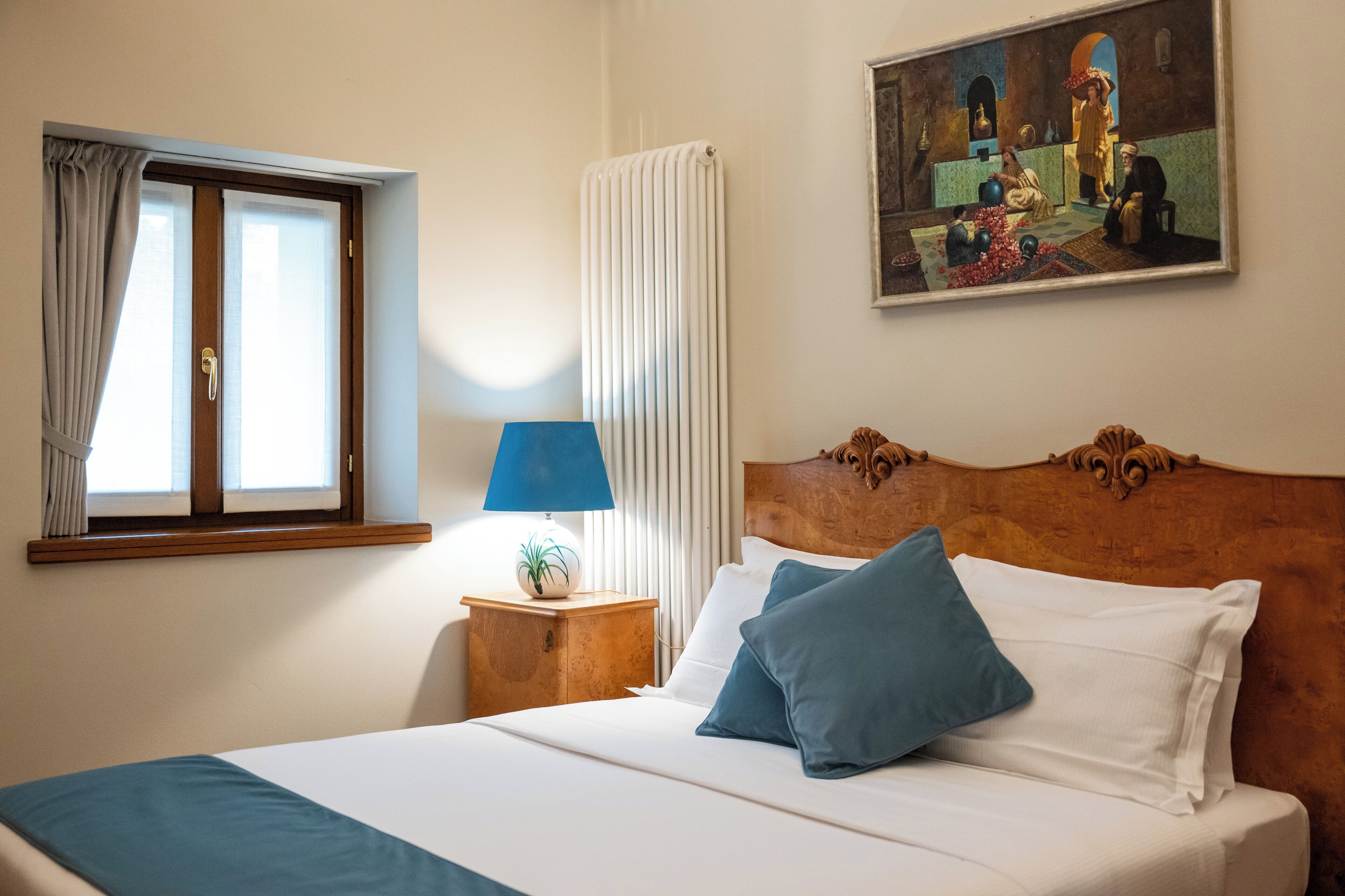 Habitación de hotel con una cama, una mesita de noche con lámpara, una ventana con cortinas y un cuadro en la pared.