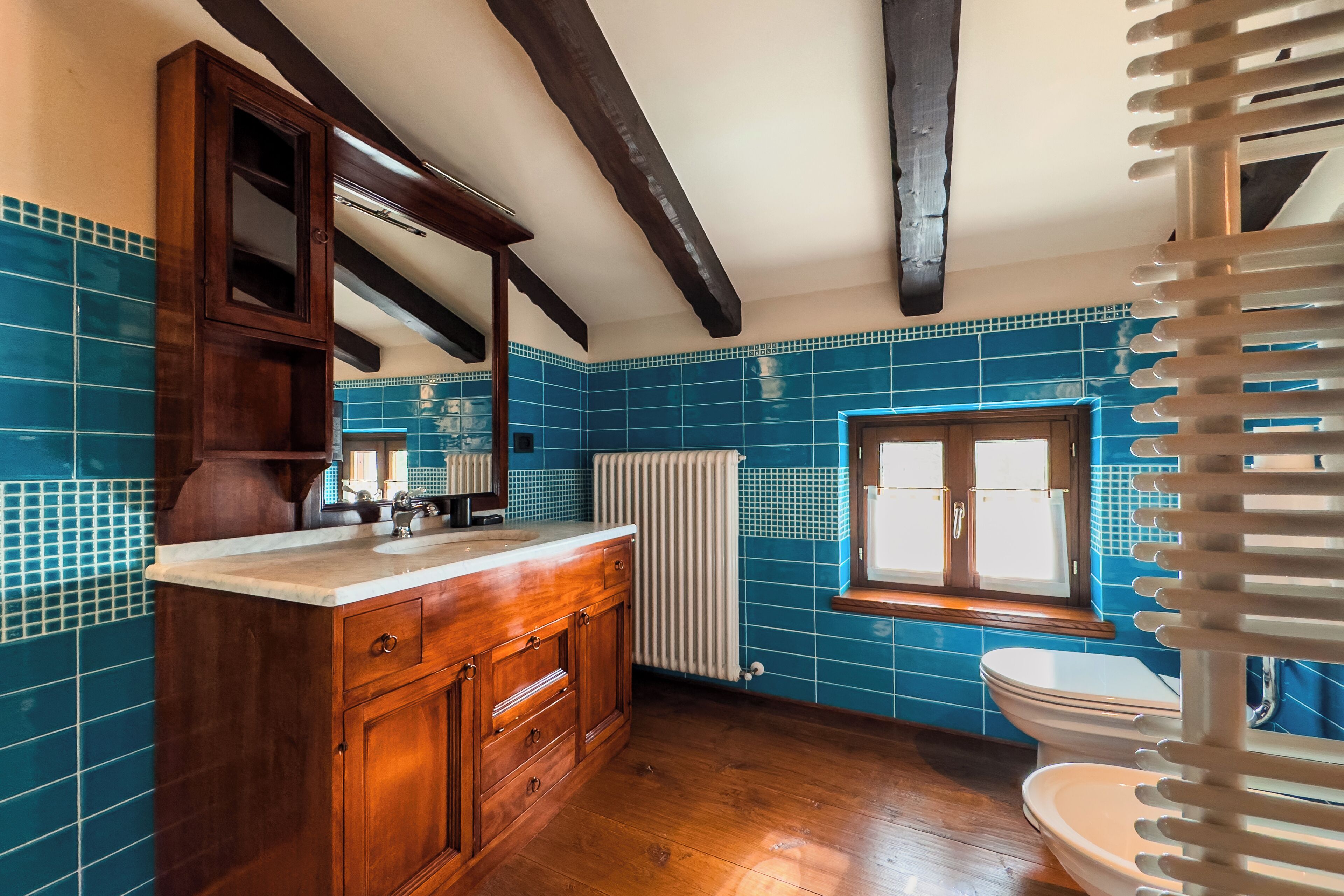 Un baño con azulejos azules, armarios de madera, un radiador blanco y una ventana.