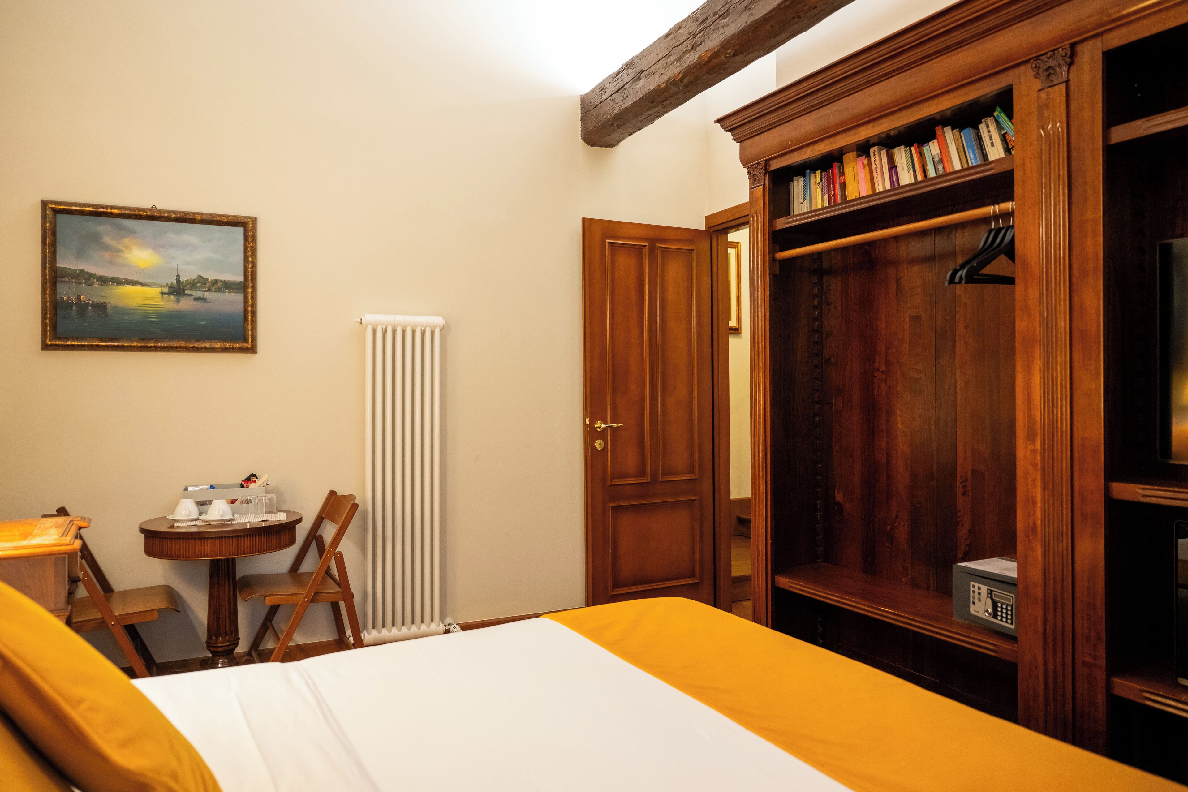 Habitación de hotel con cama, armario de madera, radiador, una mesita con silla y un cuadro en la pared.