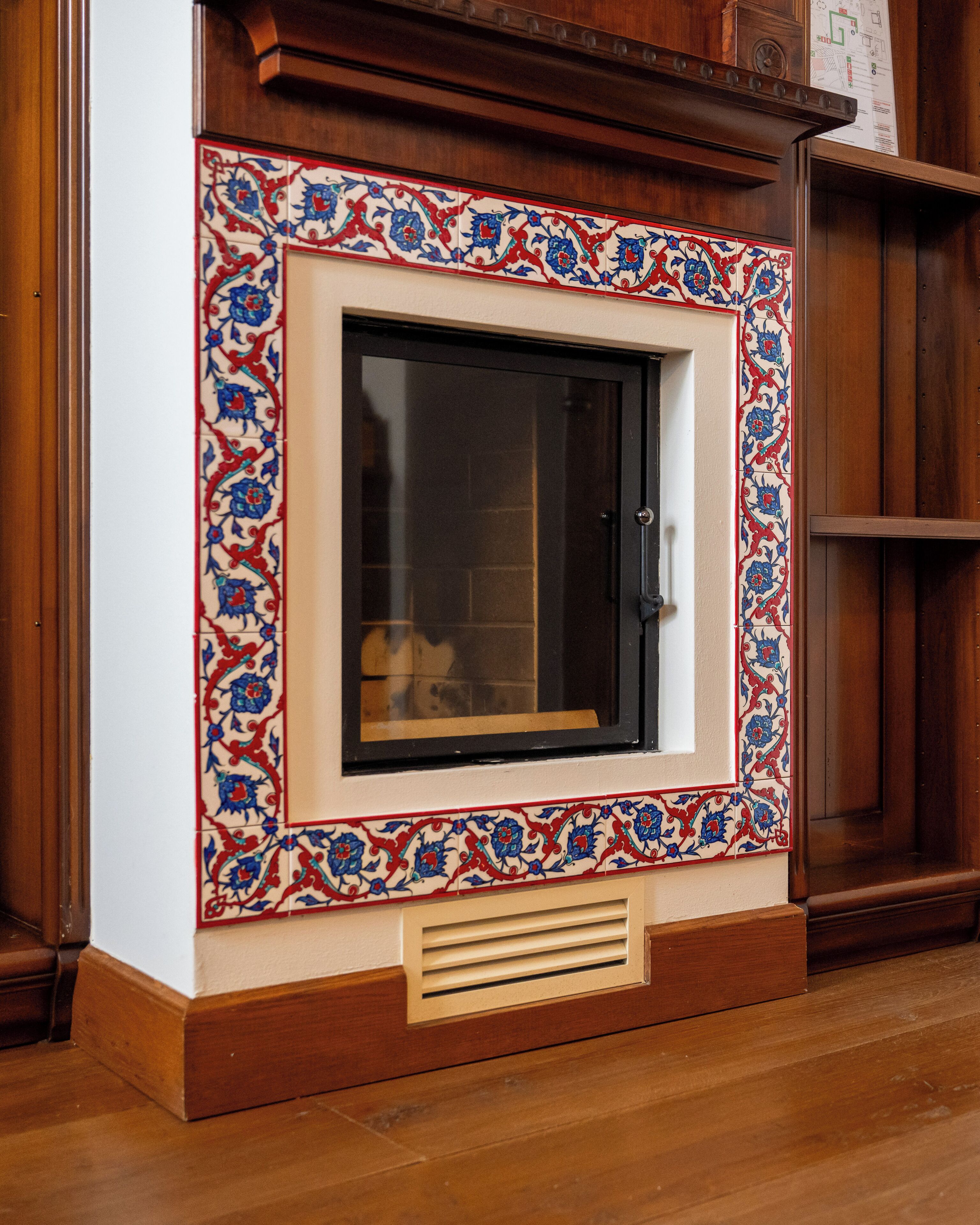 A room with a large window framed by ornate tile work, a wooden floor, and a built-in cabinet.