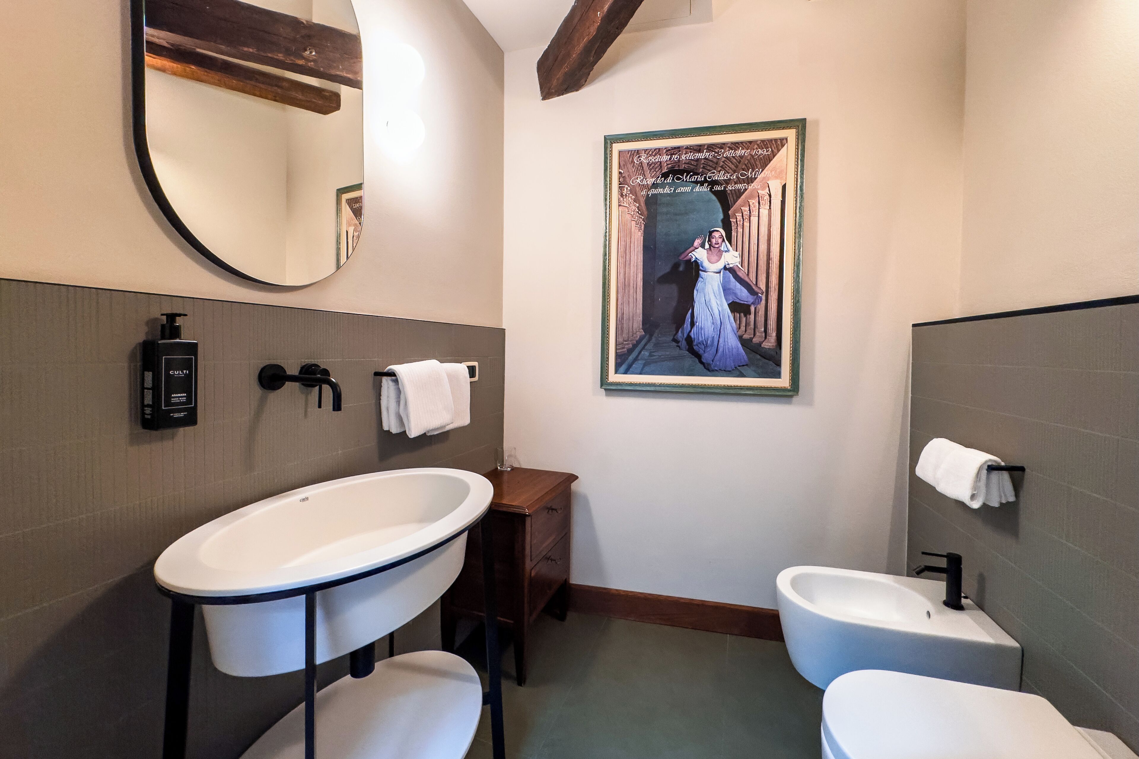 Un baño con un lavamanos ovalado grande, un mueble de tocador pedestal, un bidé y un cuadro en la pared.