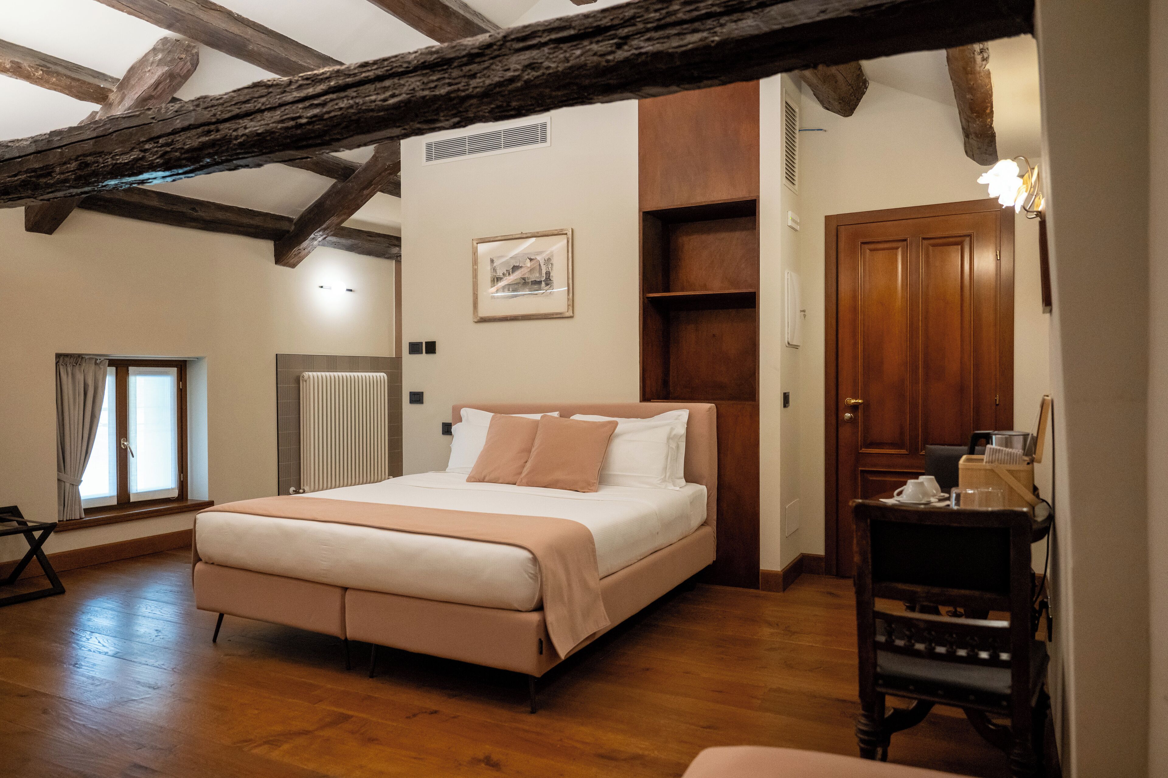Un dormitorio con una cama grande, vigas de madera, un radiador, un cuadro enmarcado, una puerta de madera y una mesita con objetos encima.
