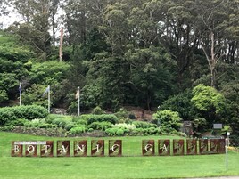 Uma placa com o texto 'BOTANICO GABRIEL' rodeada por vegetação exuberante e árvores.