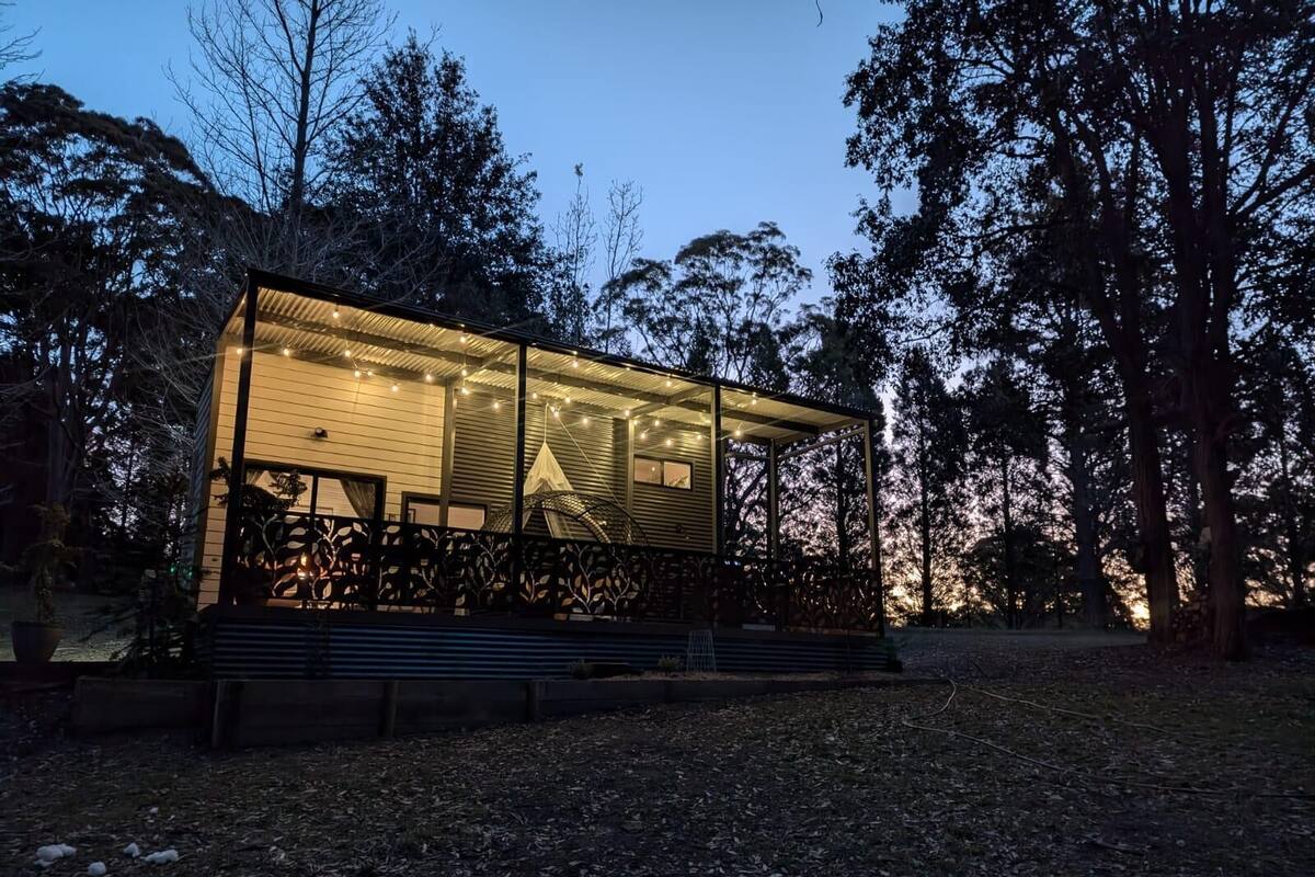 A modern, glass-walled structure with a wooden deck and a canopy, surrounded by trees.