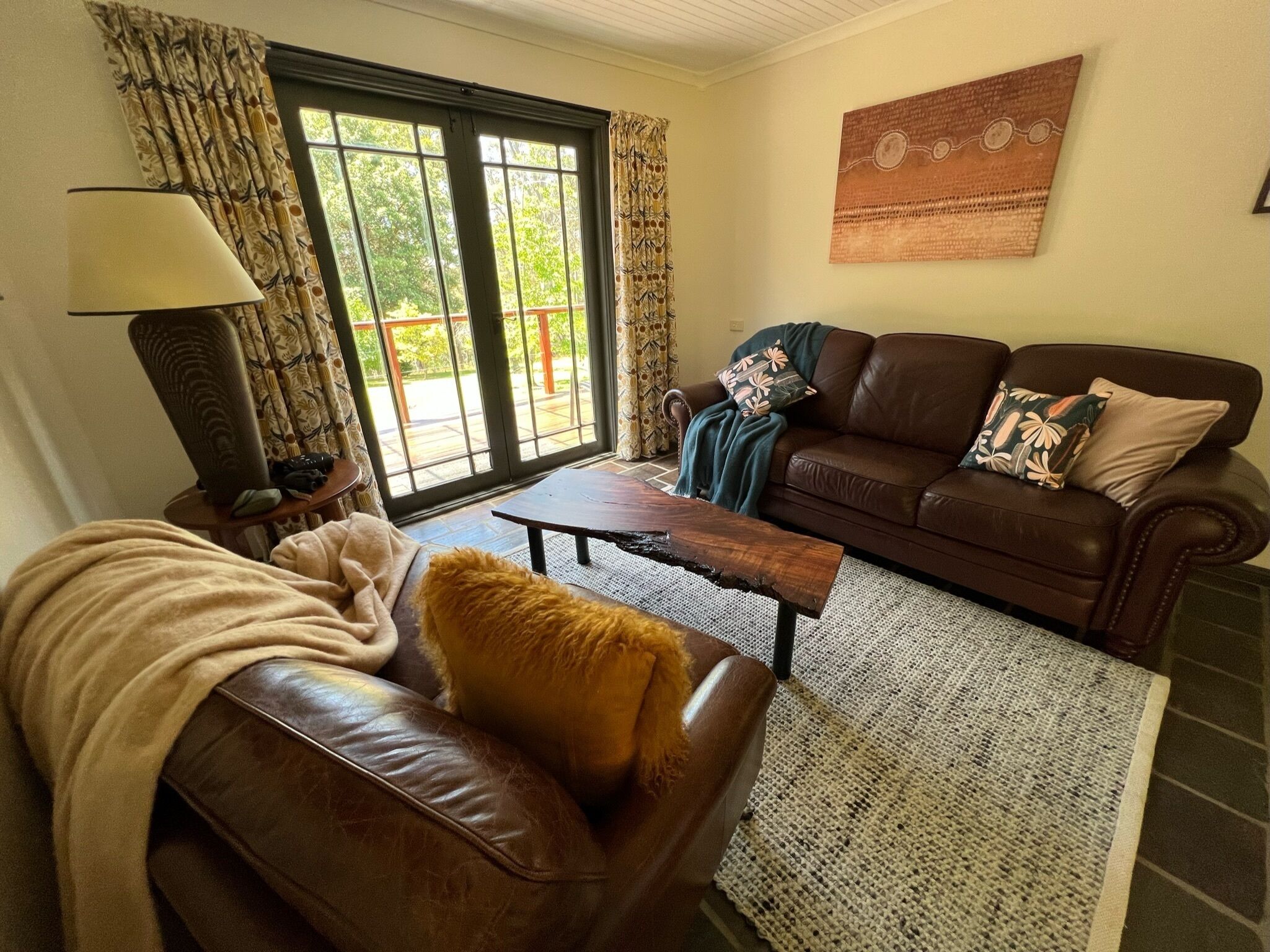 A living room with a brown leather couch, a coffee table, a lamp, and a sofa with patterned cushions. There is a large window with curtains, a rug, and a wall art piece.
