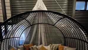 Un fauteuil suspendu circulaire, doté d’une structure en treillis et d’un auvent blanc, placé dans une pièce aux murs gris et avec une fenêtre.