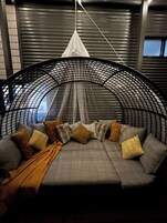 Uma cadeira suspensa circular com estrutura em tela e toldo branco, colocada em um quarto com paredes cinzas e uma janela.