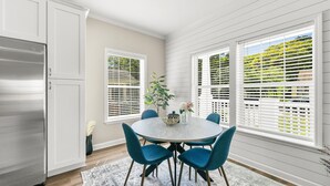 Una cocina con gabinetes blancos, un refrigerador de acero inoxidable, una mesa redonda con sillas azules y una ventana con persianas.