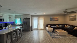 A modern kitchen with a stainless steel refrigerator, a microwave, and a sink. There is a dining area with stools and a wooden table. The living room has a dark-colored sofa and a coffee table.