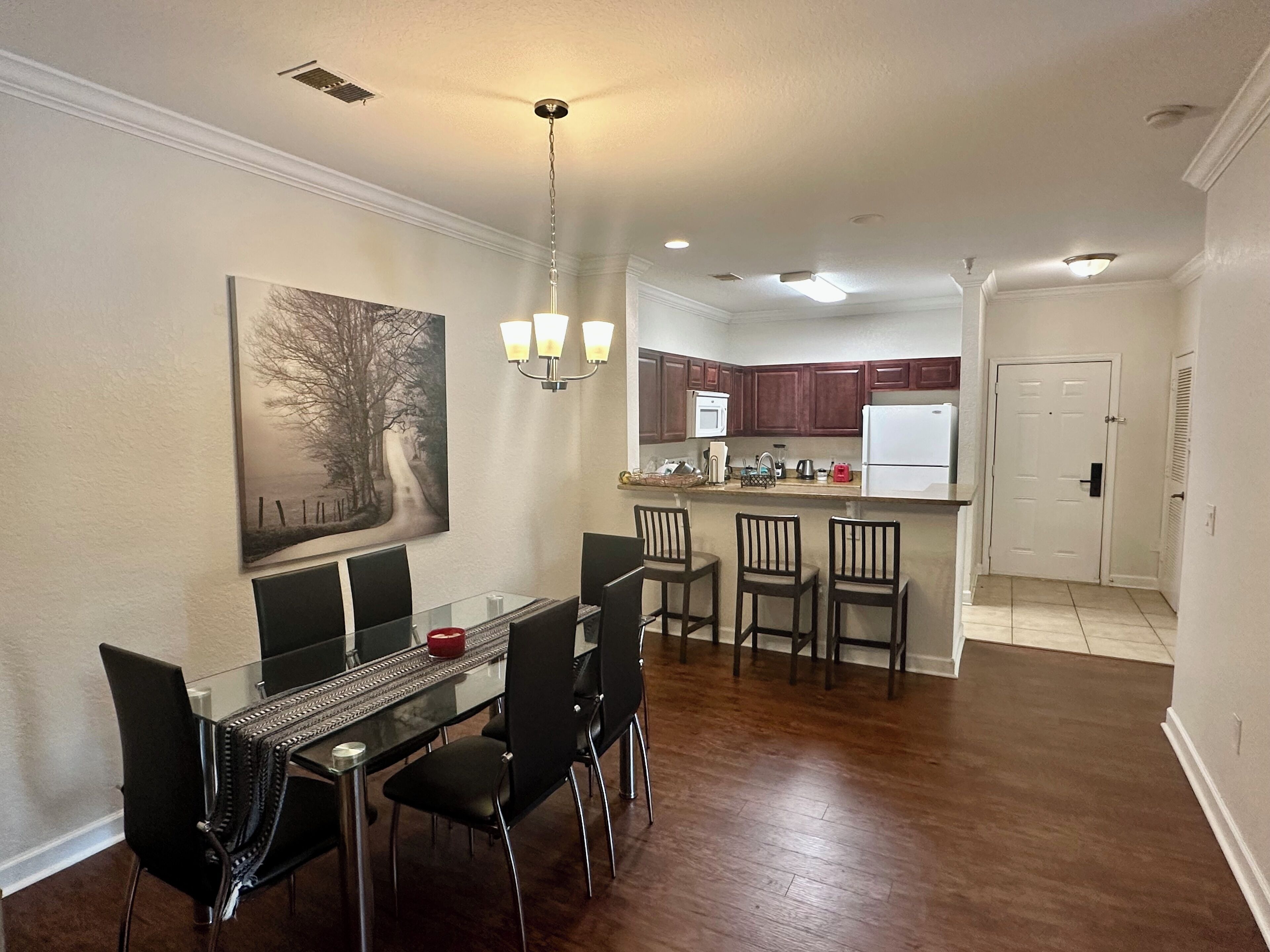 A dining area with a glass table and black chairs, a kitchen with wooden cabinets, and a door leading to another room.