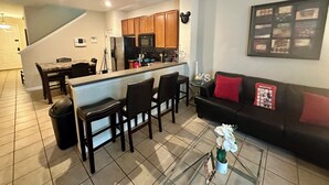 Una cocina moderna con armarios de madera, una isla central y un comedor con sillas. Se observa un sofá de cuero negro y una mesa de centro de vidrio con una orquídea en maceta.