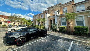Un estacionamiento con un coche deportivo negro, una señal de parada y una fila de casas adosadas color beige con detalles blancos y palmeras.