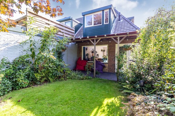 Ein kleines Haus mit Garten, Terrasse und einer überdachten Veranda mit Pflanzen.
