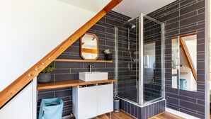 A modern bathroom with a wooden ceiling, a glass shower enclosure, a white sink, and a mirror on a black-tiled wall.