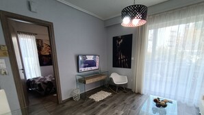 Una sala de estar moderna con una mesa de centro de vidrio, un televisor de pantalla plana sobre un mueble de vidrio, una silla blanca y un ventanal con cortinas translúcidas.
