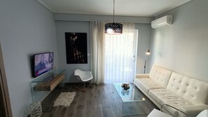 Una sala de estar moderna con un sofá, una mesa de centro de vidrio, un televisor de pantalla plana, una silla blanca y un ventanal con cortinas translúcidas.