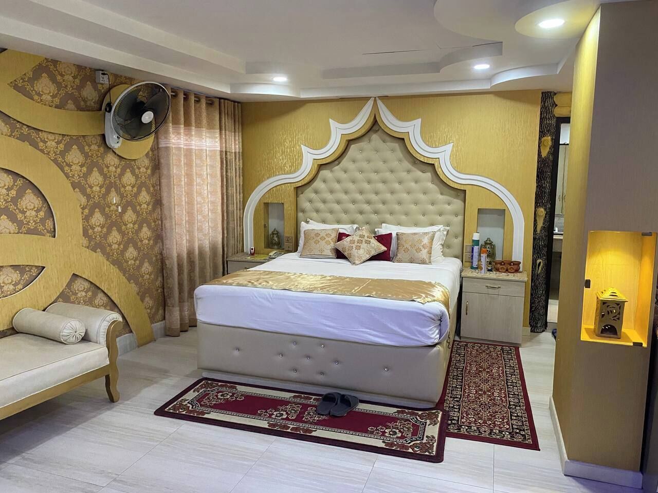 A bedroom with a large bed, a bedside table, a nightstand, a wall-mounted fan, and a decorative wall panel.
