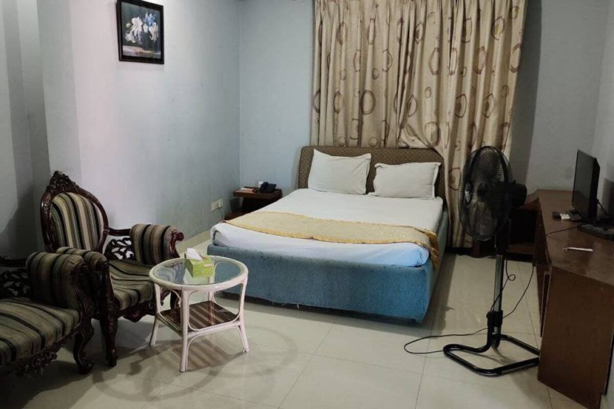 A hotel room with a bed, two chairs, a small table, a desk, a fan, and a clock on the wall.