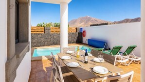 Área de piscina com mesa posta para refeições, cadeiras de lounge e uma parede de pedra.