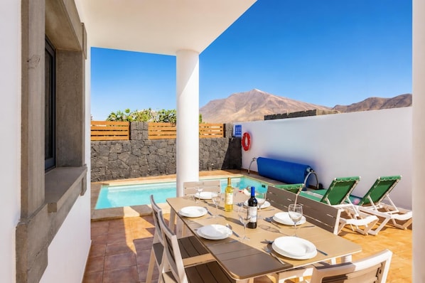 Área de piscina com mesa posta para refeições, cadeiras de lounge e uma parede de pedra.