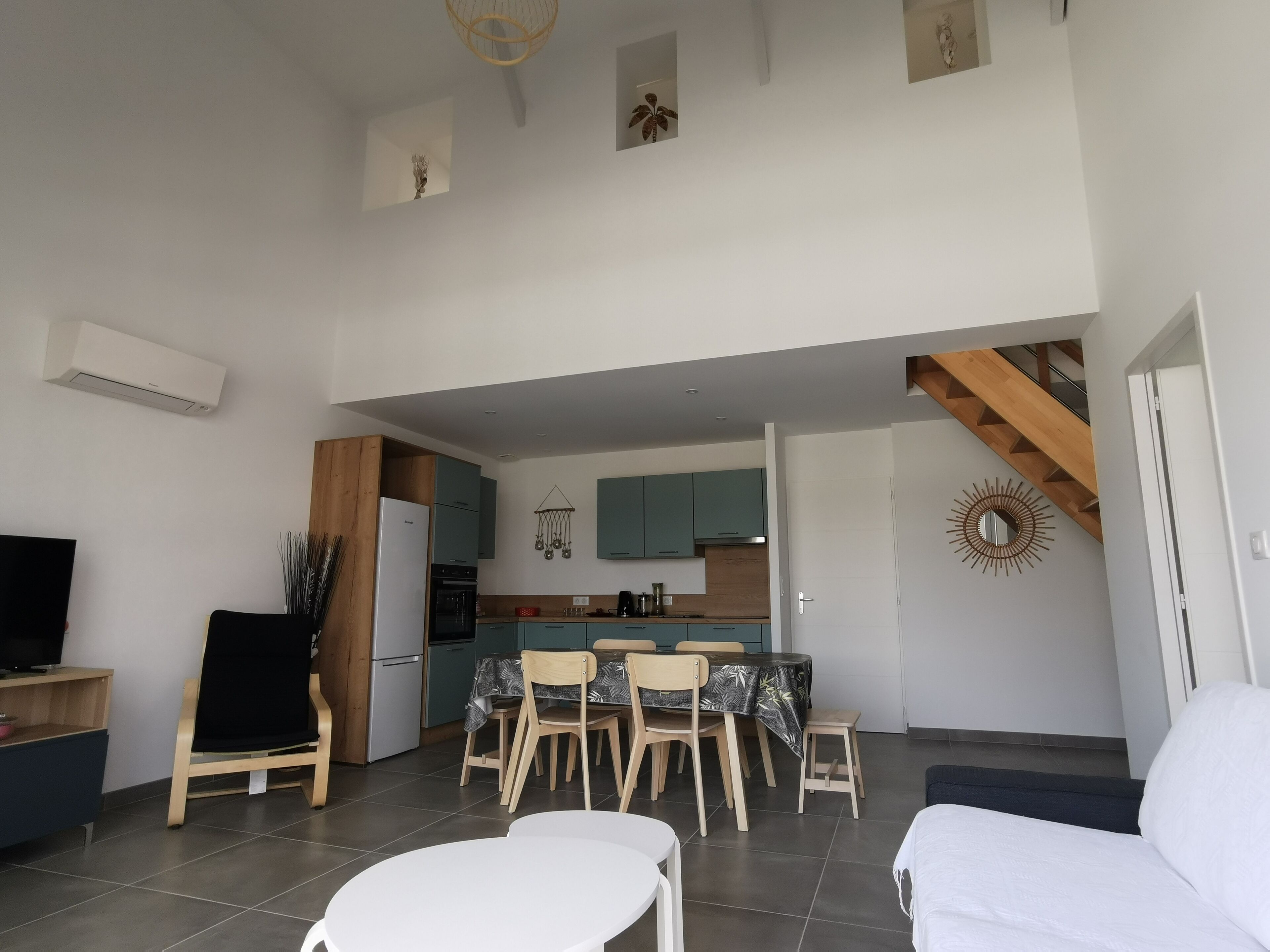 A modern living room with a sofa, a dining table with chairs, a kitchen area with cabinets and a stove, and a staircase leading to another level.