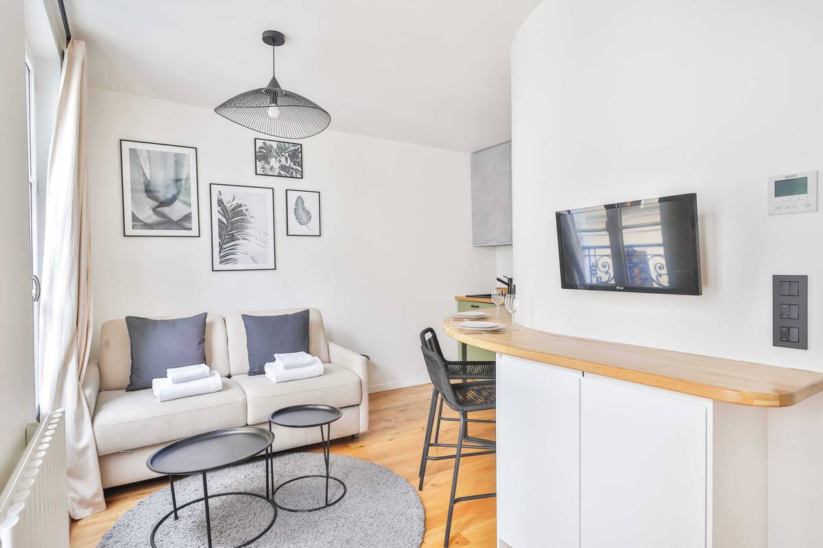 A modern living room with a sofa, a small round table, and a dining area with a wooden counter and chairs.