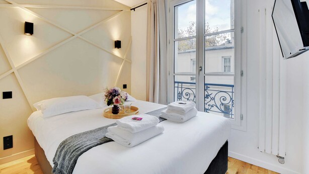 Ein ordentlich bezogenes Bett mit weißen Leinen, ein Nachttisch mit einer Blumenvase und ein Fenster mit Blick nach draußen.