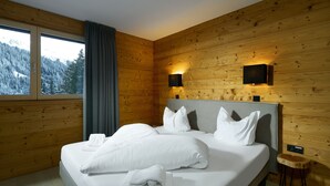 Una habitación con paneles de madera, una cama, dos luces fijadas a la pared y un ventanal con vistas a árboles nevados.