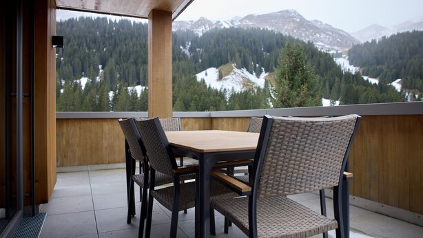 A balcony with a wooden table and wicker chairs, overlooking a mountainous landscape with snow patches and dense forests. - 59111 Bartgeier 111 T (Gargellen)
