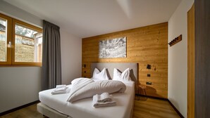 A neatly made bed with white linens and pillows, a wooden headboard, a window with curtains, and a wooden floor.
