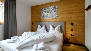 Un lit bien fait, avec du linge de lit blanc et des oreillers, un mur en bois orné d’un tableau, et un petit tabouret en bois.